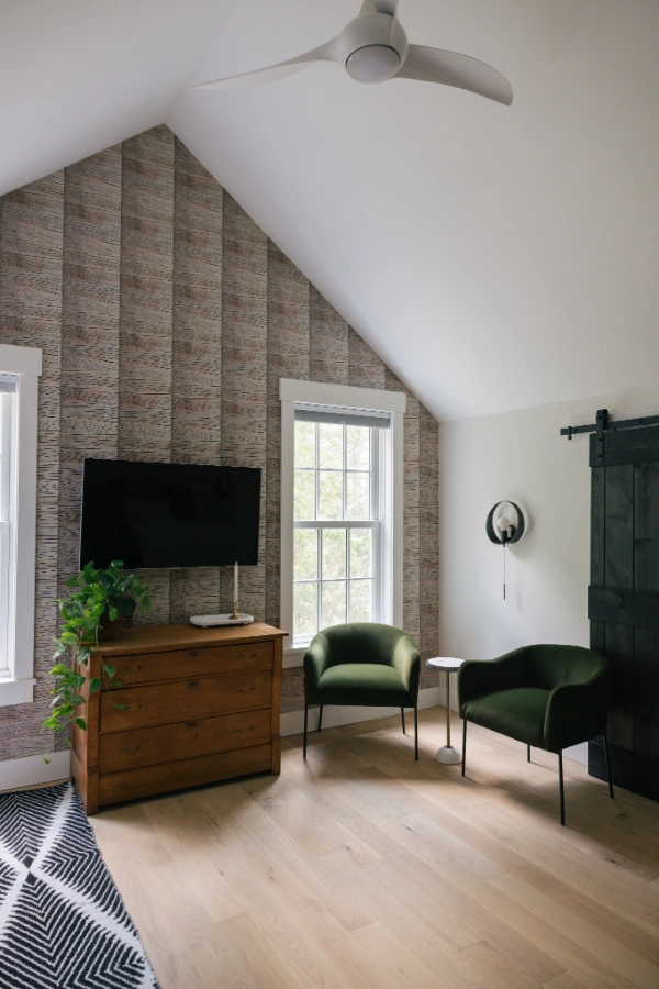Vaulted upstairs bedroom with textured accent wall, mounted television above wood dresser, and pair of green lounge chairs beneath ceiling fan